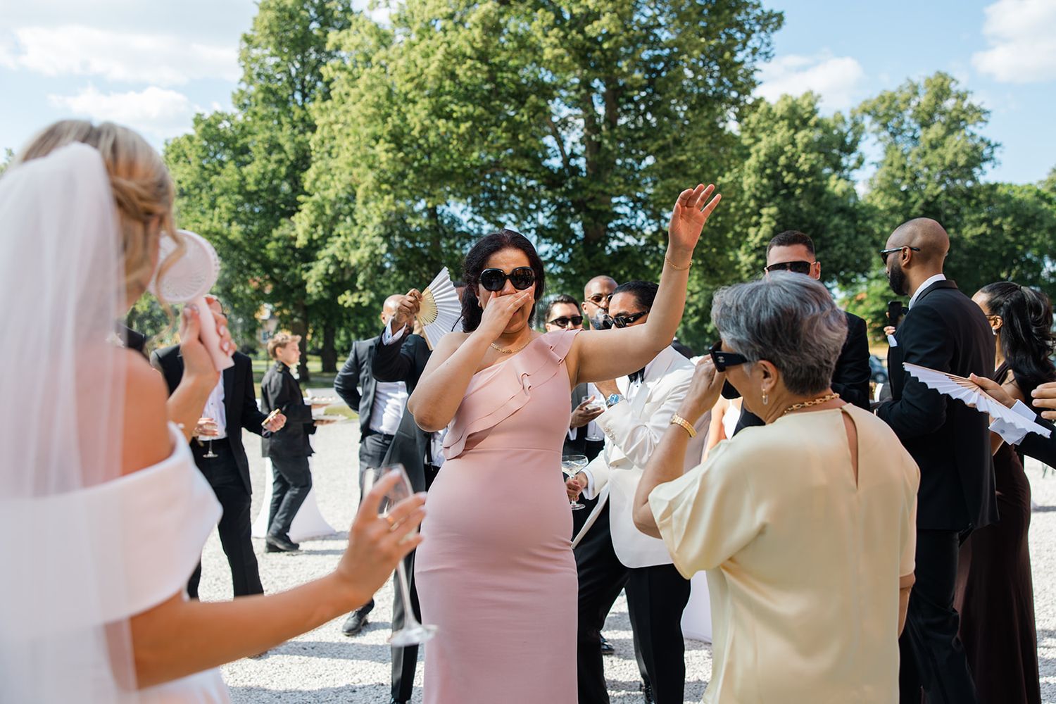 Wedding guests celebrate and wave at an outdoor reception on a sunny day with trees in the background.