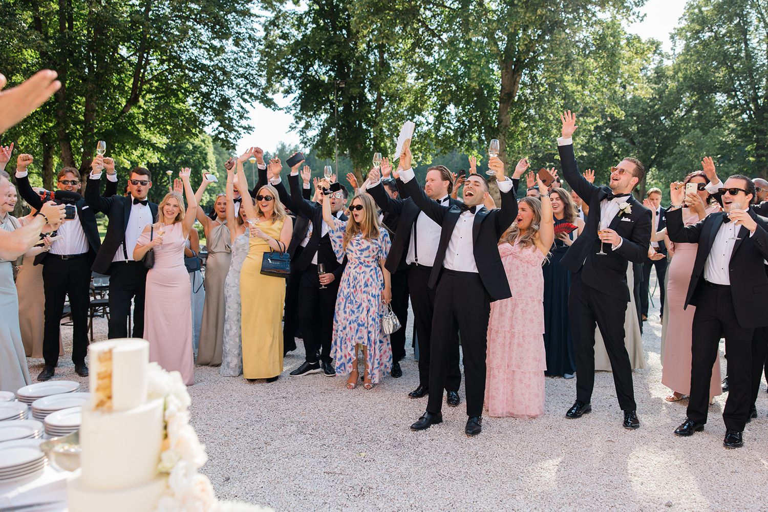 Wedding guests and bridal party celebrate during an outdoor reception with raised arms and joyful expressions.