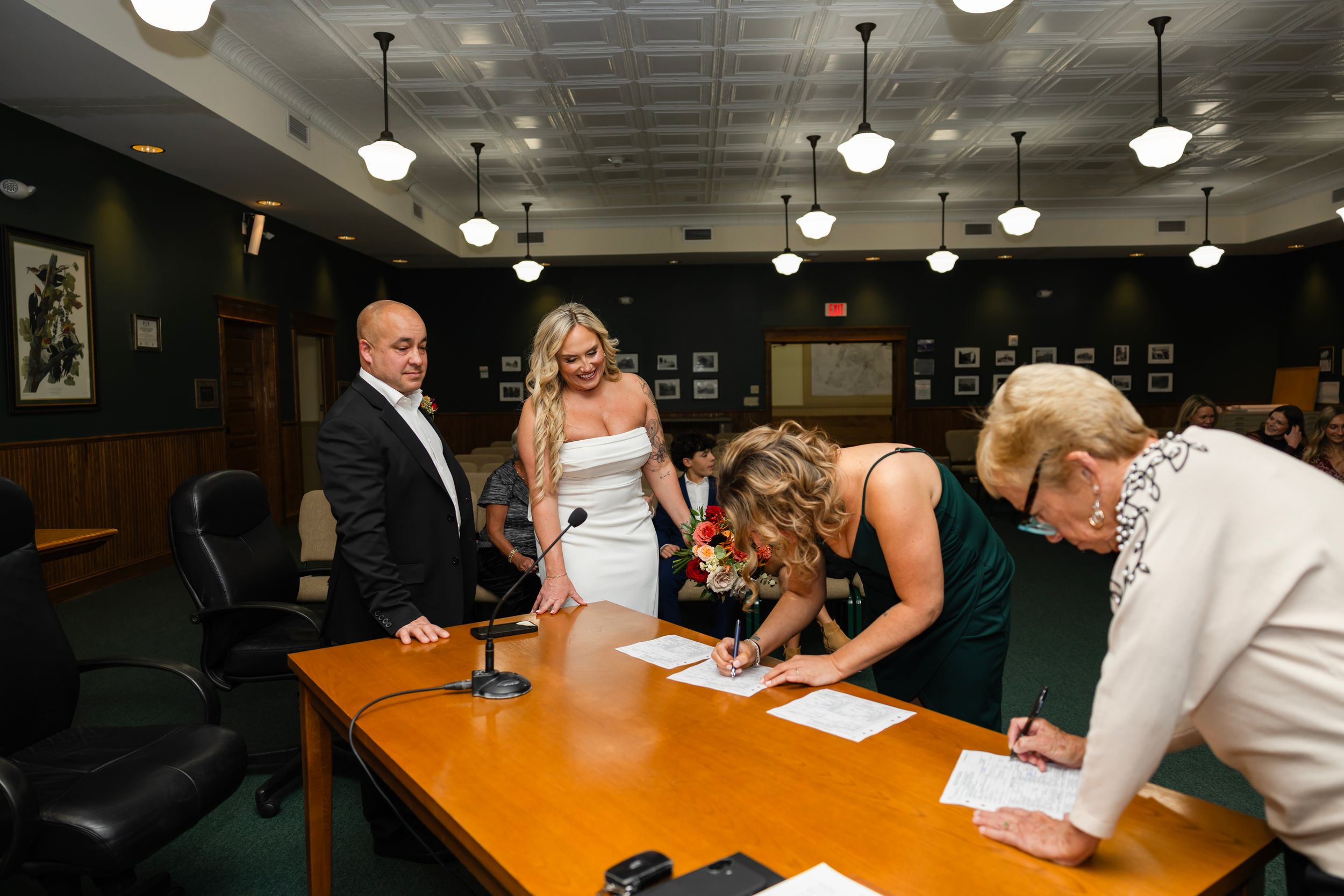 Wedding ceremony paperwork being signed at a long wooden table with multiple witnesses present.