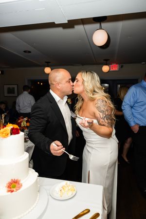 A newlywed couple shares a romantic kiss next to their white wedding cake decorated with orange and yellow flowers | NJ wedding photographer