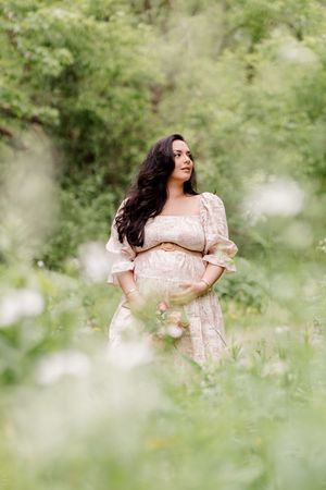 Future maman dans une robe rose et blanche entourée de verdure et de fleurs dans un boisé près des rives du fleuve Saint-Laurent