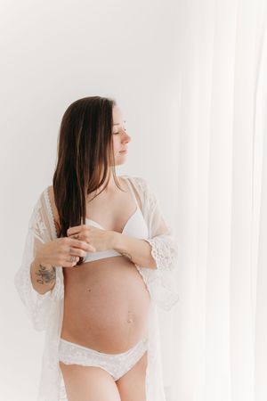 Portrait artistique de grossesse d’une maman en sous-vêtements de dentelle coordonnés qui porte une main à sa chevelure