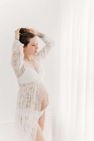 Profil fine art d’une future maman dans une robe de dentelle diaphane qui remonte ses cheveux en un chignon sur sa tête au studio de Québec