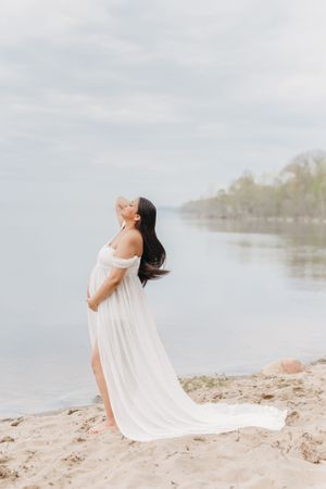 Future maman dans une robe de voilages blancs sur une plage de sable au bord du fleuve Saint-Laurent à Saint-Augustin