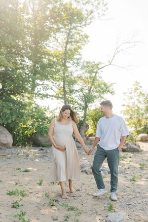 Couple de futurs parents se souriant main dans la main en avançant sur une plage bordée de grands arbres dans le soleil matinal