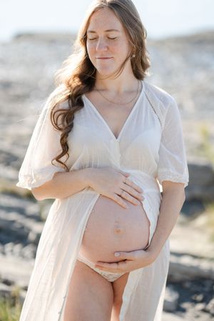 Future maman dans une robe de coton léger au bord du fleuve à Neuville tenant amoureusement son ventre les yeux fermés