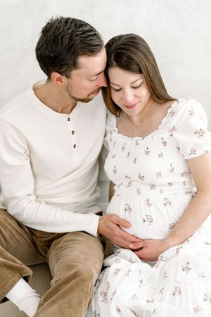 Futurs parents enlacés qui baissent les yeux vers le ventre de la future maman habillée d’une robe blanche de la garde-robe cliente