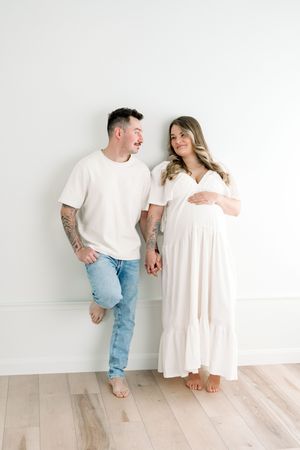 Portrait de futurs parents adossés contre un mur blanc se tenant la main en se regardant au studio de photographie de Québec