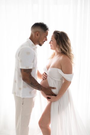 Couples de futurs parents en contre-jours dans des vêtements blancs se regardant avec tendresse au studio près de Québec