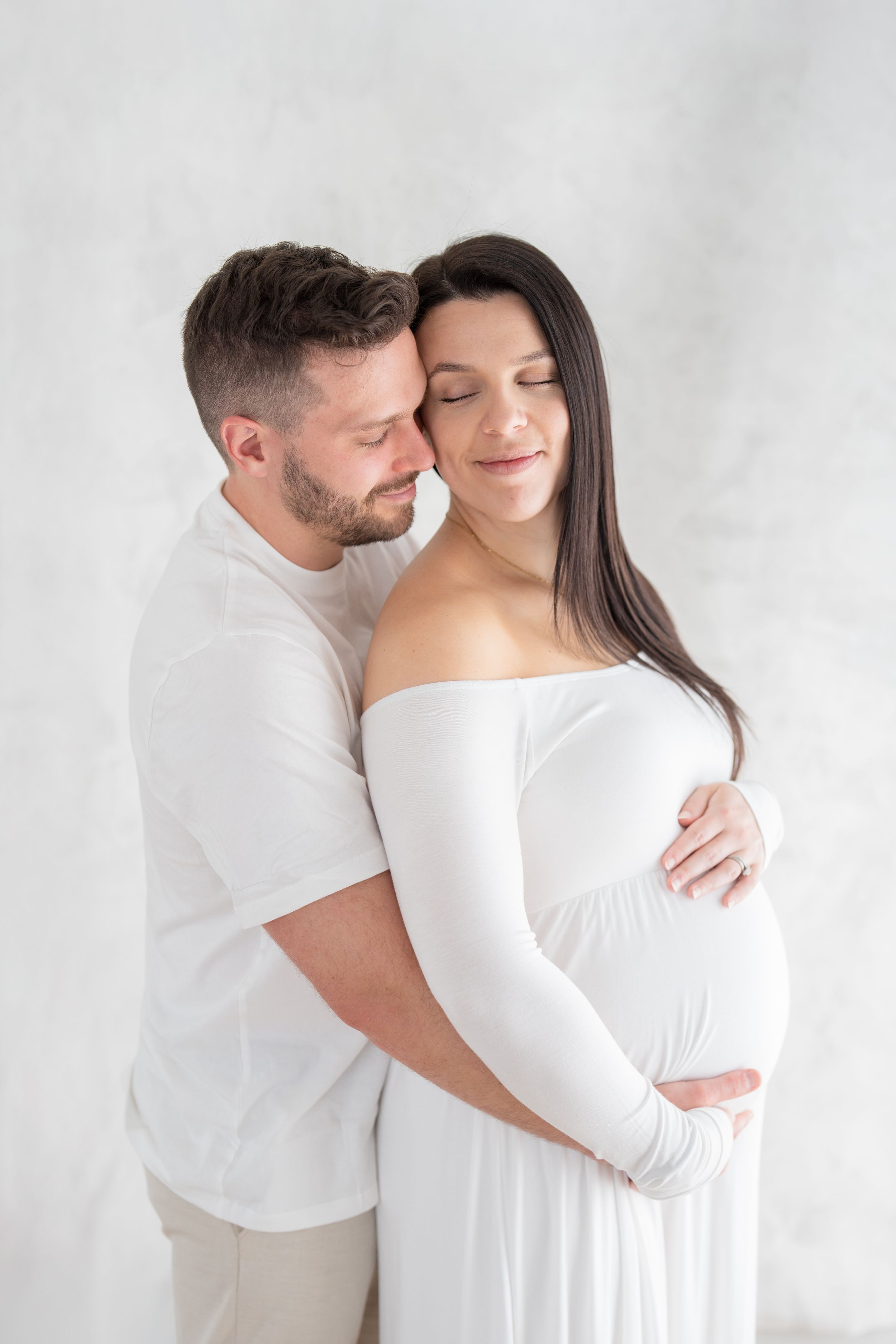 Futurs parents dans un moment d'émotion tendre habillés de blanc lors de leur séance photo de maternité fine art au studio de Québec
