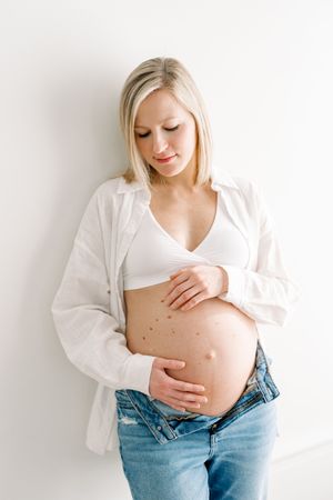 Future maman habillée d’une chemise blanche ou ouverte et d’un jeans portant les mains à son ventre lors de sa séance fine art maternité