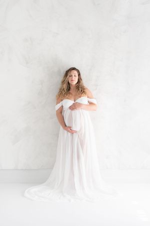 Future maman dans une robe de voilages blancs vaporeuse contre une toile fine art peinte à la main lors d'une séance photo de grossesse
