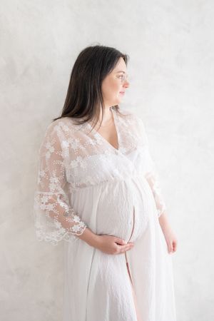 Portrait d’art de grossesse d’une maman qui regarde au loin en plaçant une main sous son ventre dans une robe bimatière de dentelle