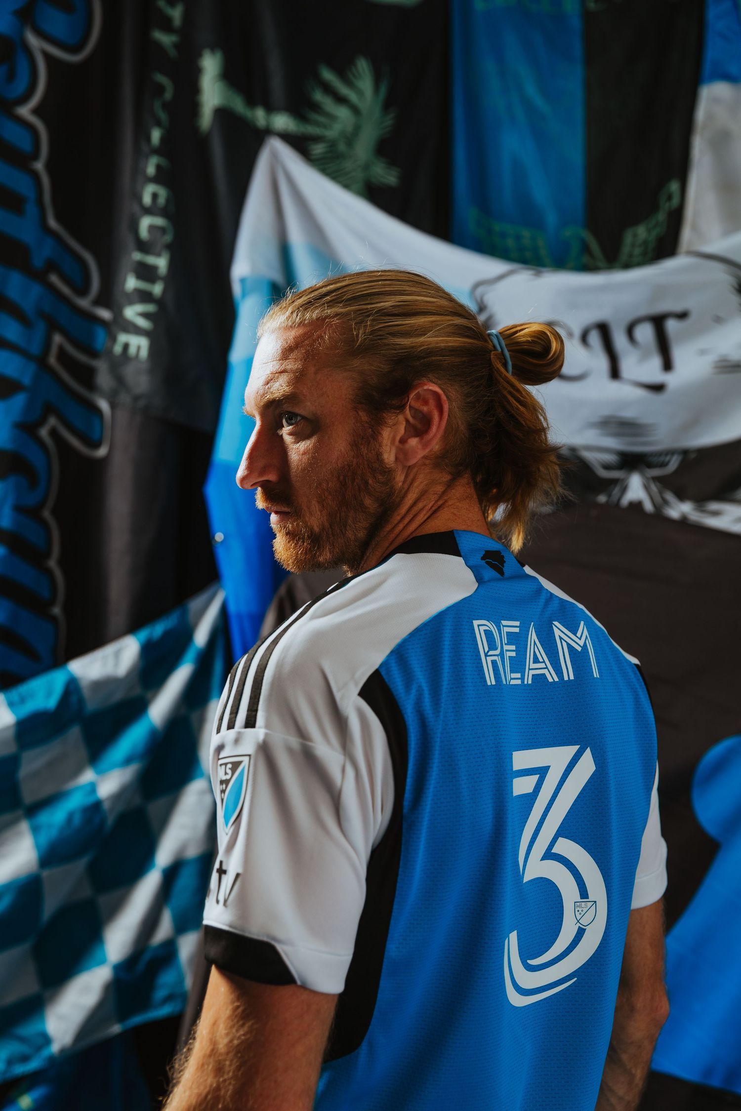 Charlotte FC player Ream in a blue Adidas number 3 jersey poses in profile before supporter flags.