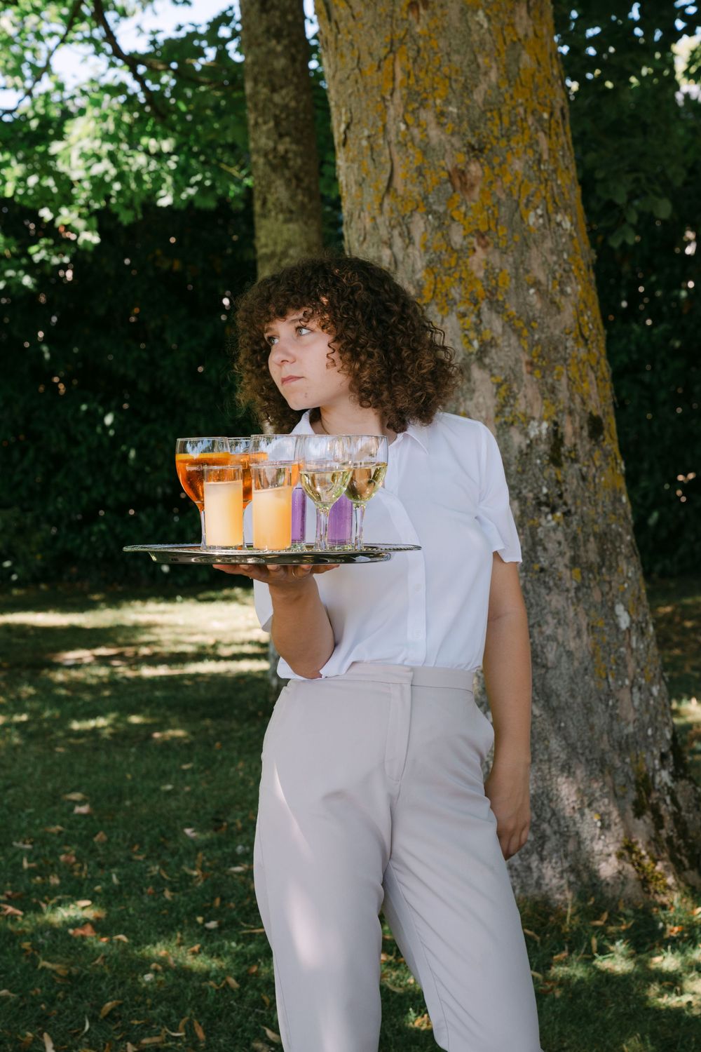 Server in white outfit carries colorful drinks on a tray in an outdoor garden setting.