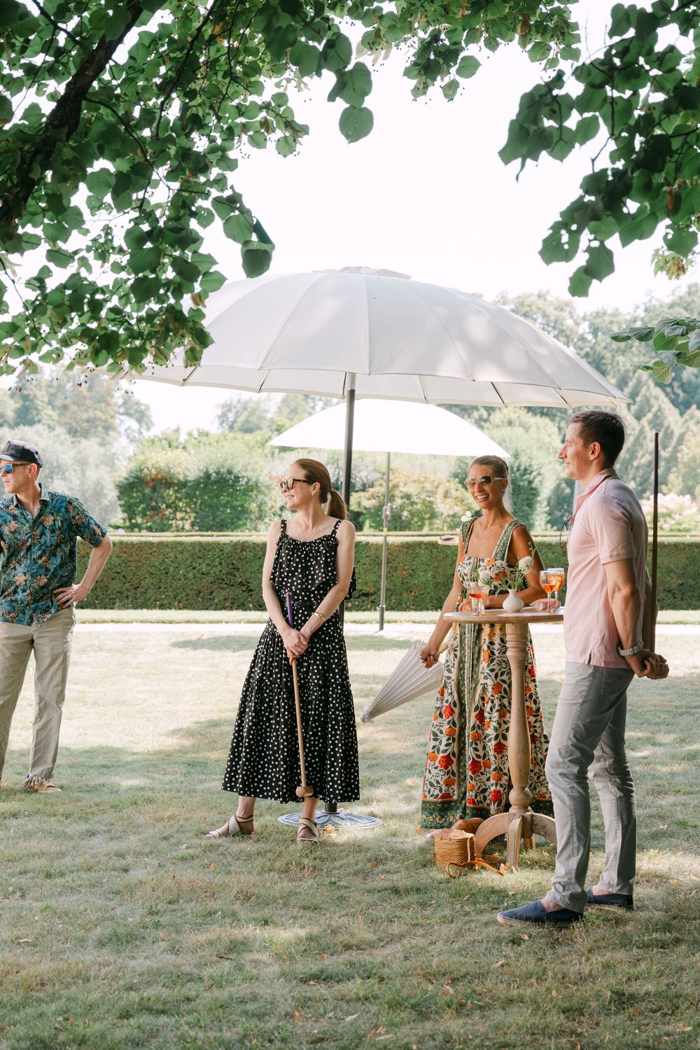 Wedding guests with umbrellas gather on a grassy lawn during an outdoor summer celebration.