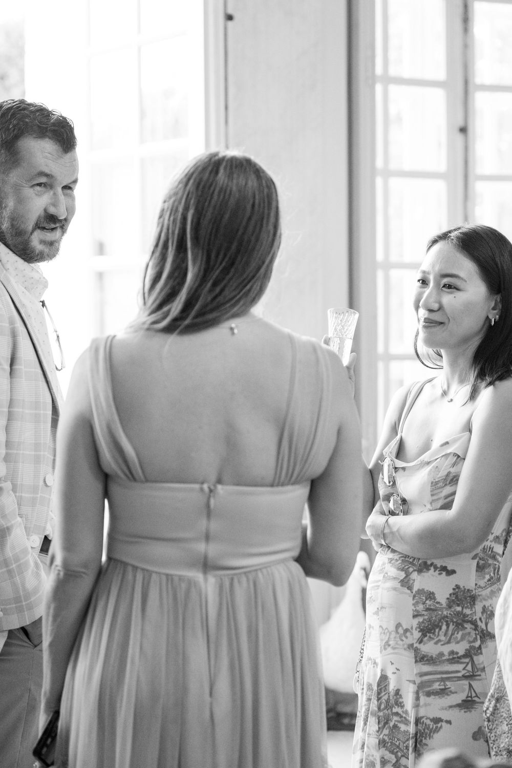 Wedding party members in light colored dresses and formal wear gathered in a bright sunlit room.