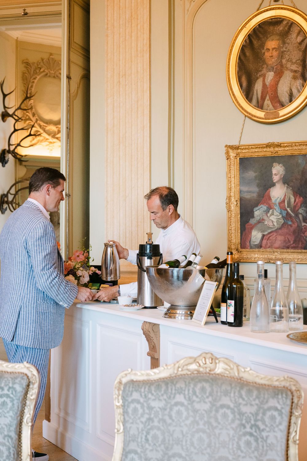 Formal dining room with ornate gold-framed portraits and staff preparing a beverage service.