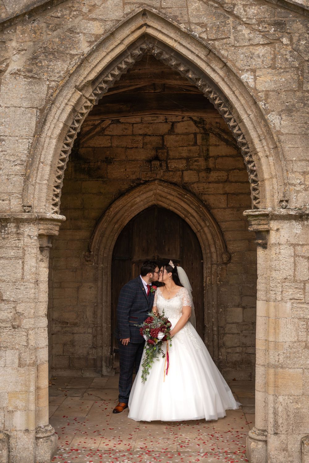 A romantic wedding couple poses under a series of historic stone archways in an ancient church or castle setting.