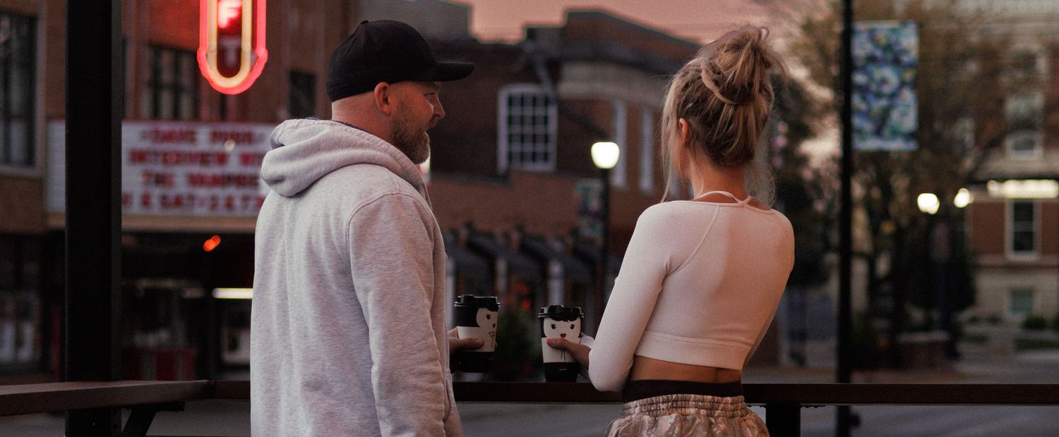 A man and woman share a coffee at sunrise, an old theater is visible in the background