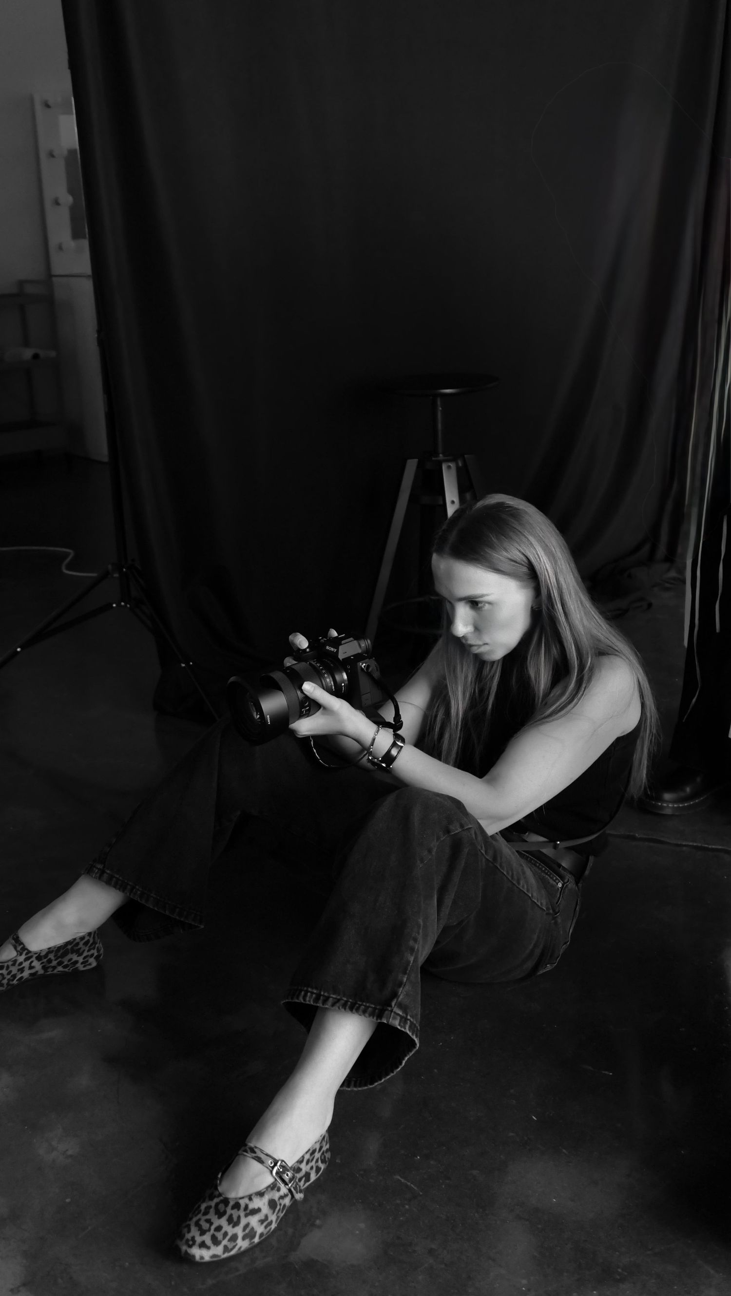 Black and white photo of photographer sitting on floor with camera against dark backdrop in studio setting.