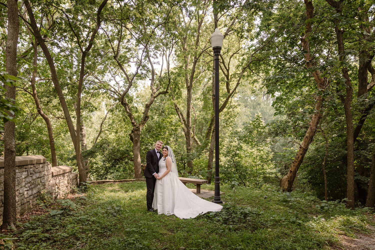 A couple shares a romantic moment in a lush green garden path surrounded by tall trees and natural light.