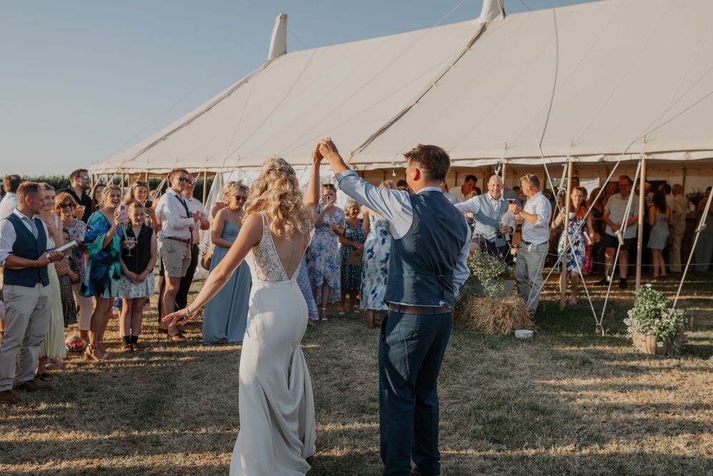 Bride and groom make grand entrance to cheering wedding guests outside a white sailcloth marquee tent.