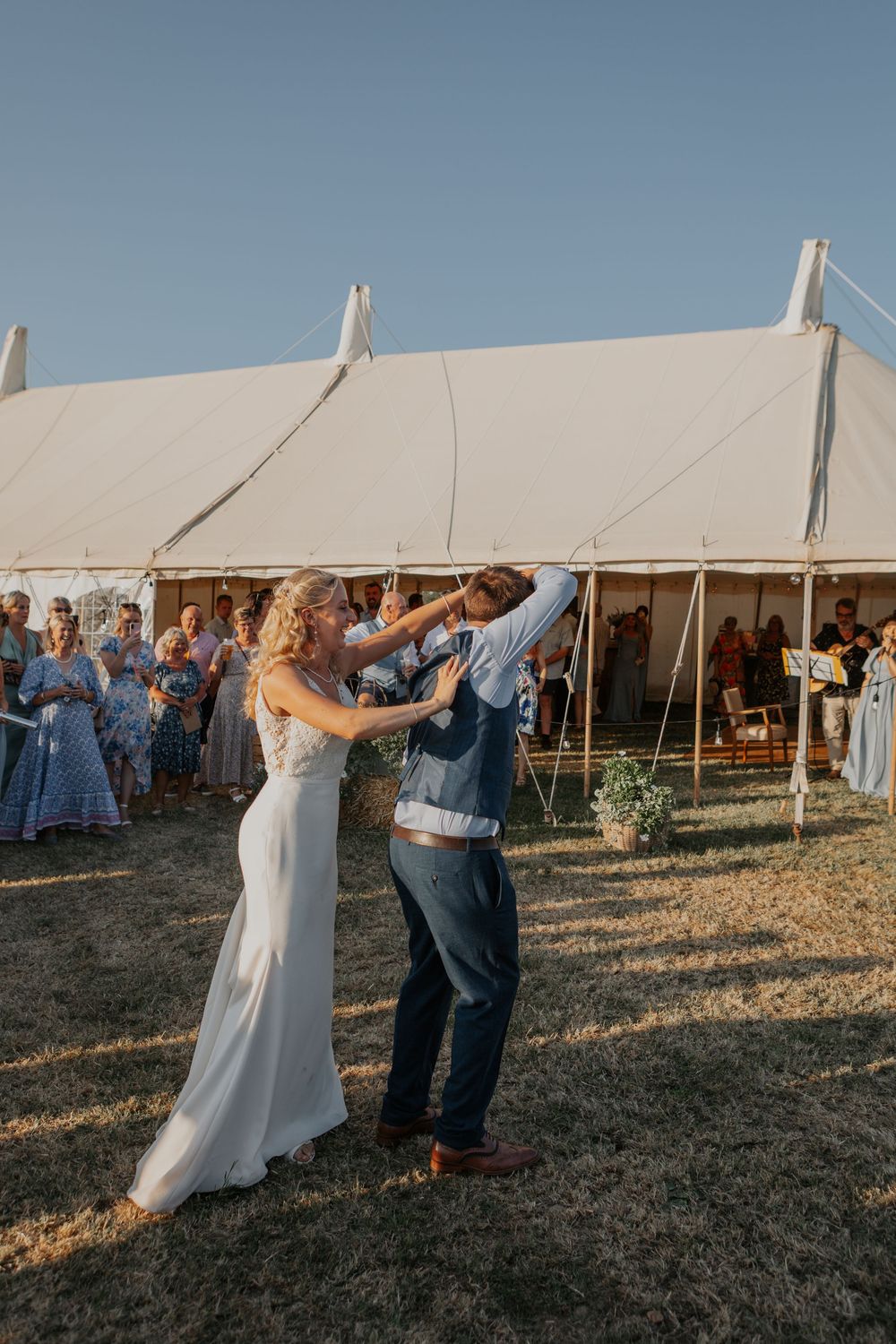 Bride and groom dance joyfully outdoors at golden hour with wedding guests watching beside a white tent.