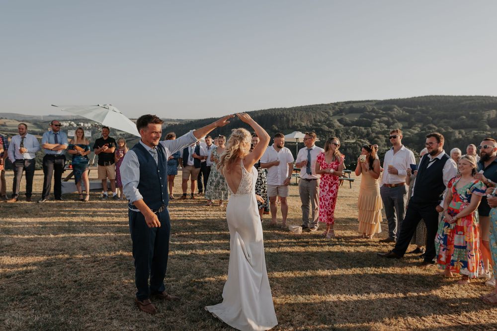 Smiling bride twirls during outdoor first dance while wedding guests cheer in a scenic countryside setting.