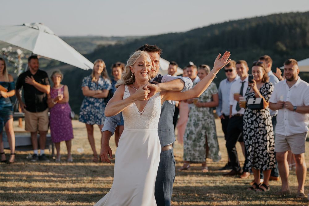 Radiant bride laughs during outdoor first dance with groom as guests celebrate in warm evening sunlight.