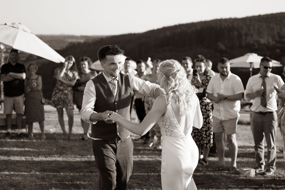 Black and white photo of bride and groom sharing first dance outdoors with guests and rolling hills behind.