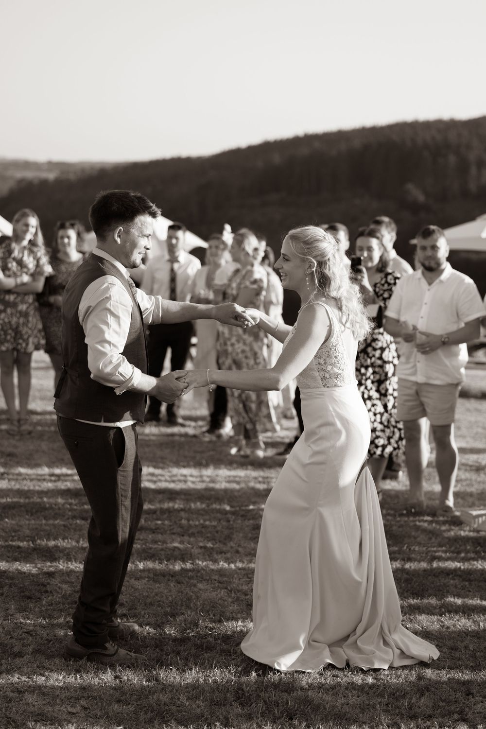 Black and white photo of newlyweds dancing outdoors as wedding guests watch in a scenic countryside setting.