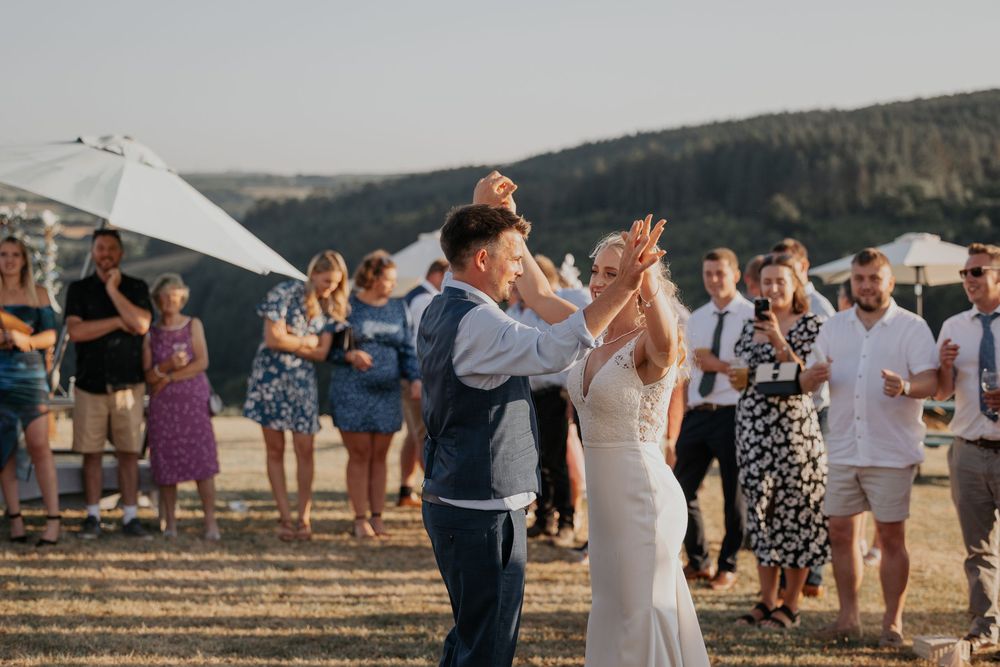 Groom lifts bride during joyful outdoor first dance as guests clap and cheer in golden evening light.