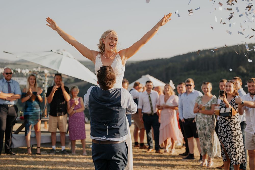 Bride lifted by groom with arms outstretched as confetti flies at an outdoor countryside wedding celebration.