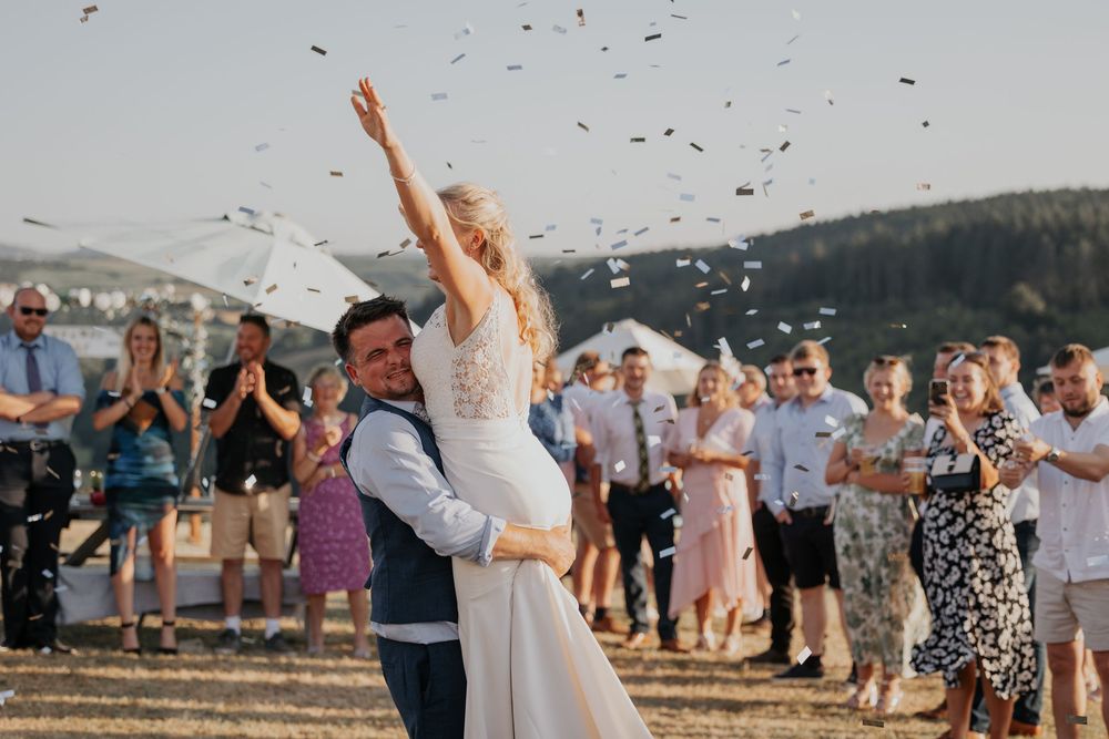 A groom lifts his bride as confetti falls around them at an outdoor wedding celebration.