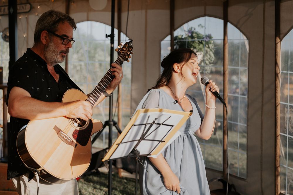 A man plays acoustic guitar while a young girl sings into a microphone at a wedding reception.