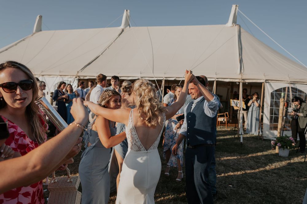 Wedding guests celebrate outside a large white marquee tent as the bride and groom walk through.