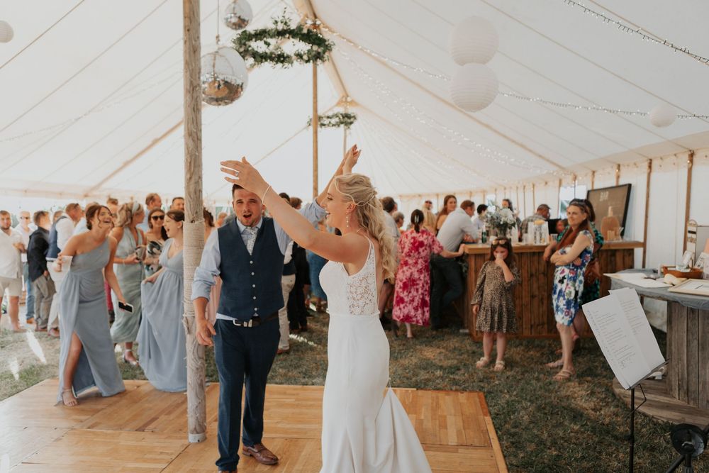 A bride raises her arm joyfully while dancing with her groom inside a sunlit wedding marquee.