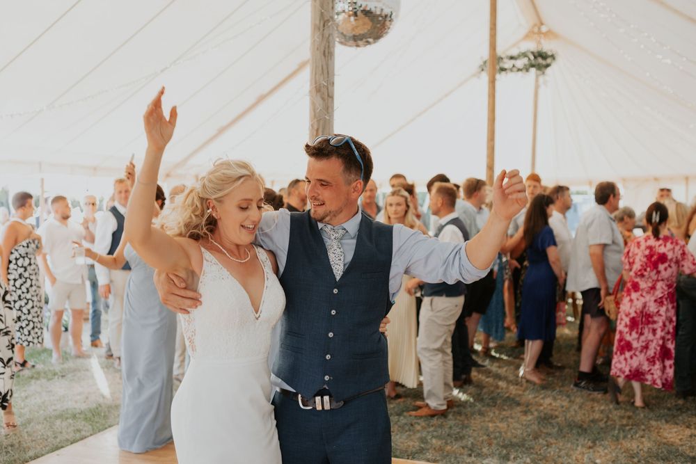 A smiling bride and groom dance together with raised hands inside a festive wedding marquee.