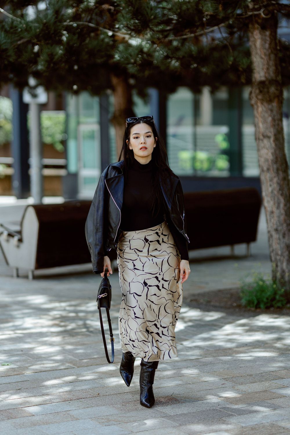 A person wearing a black top and patterned skirt walks along a tree-lined path in an urban setting.