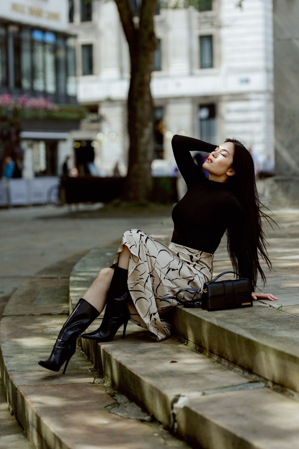 Fashion model in black outfit and boots poses gracefully on concrete steps in urban setting.