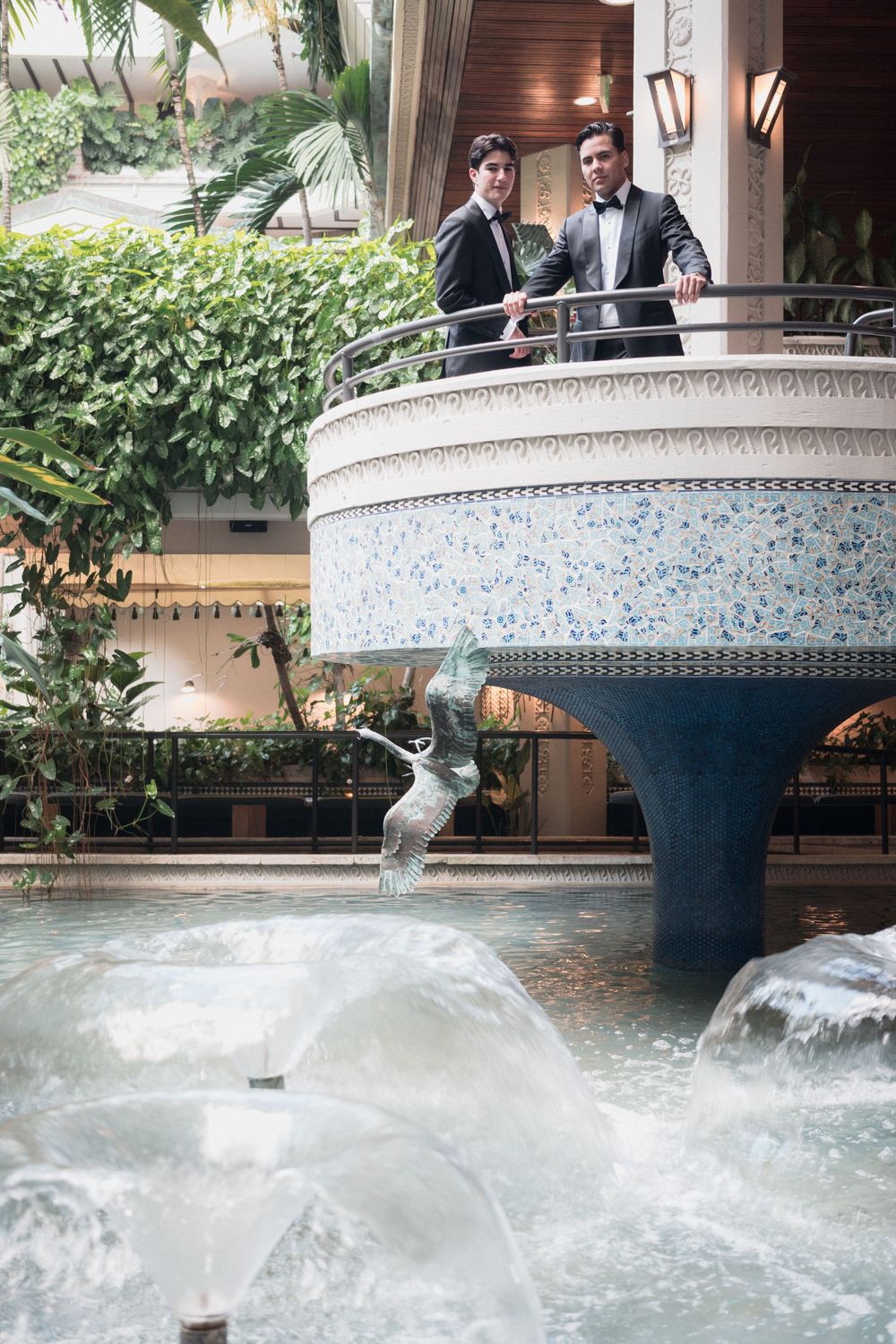 Two elegantly dressed individuals stand on a decorative balcony overlooking an ornate fountain in a lush indoor atrium.