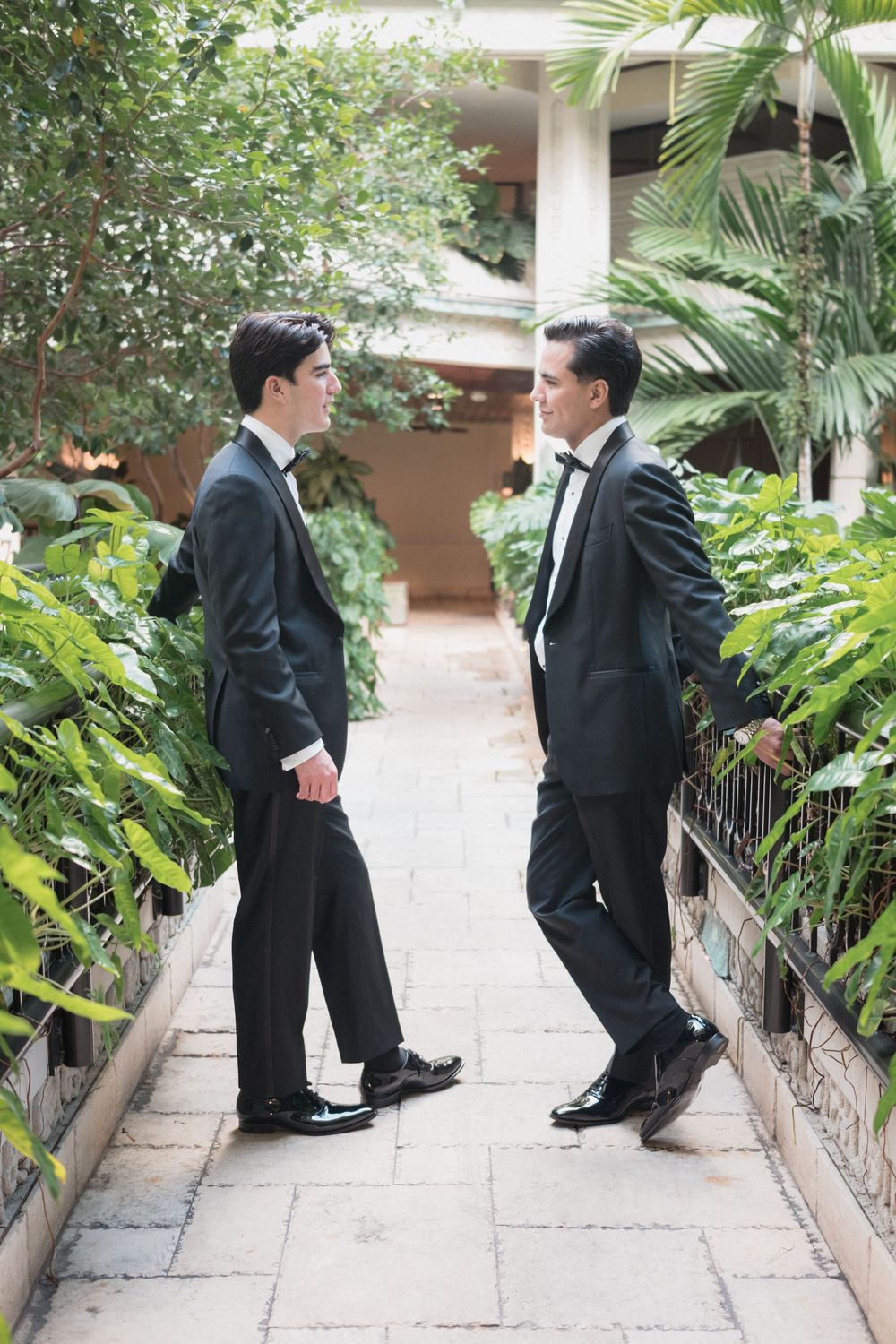 Two men in black tuxedos face each other on a garden pathway surrounded by lush green tropical plants.
