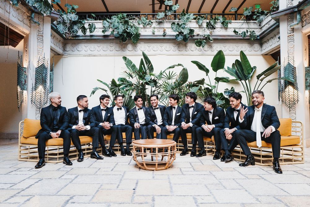 Groomsmen in black tuxedos sit together on wooden benches in an elegant indoor courtyard setting.