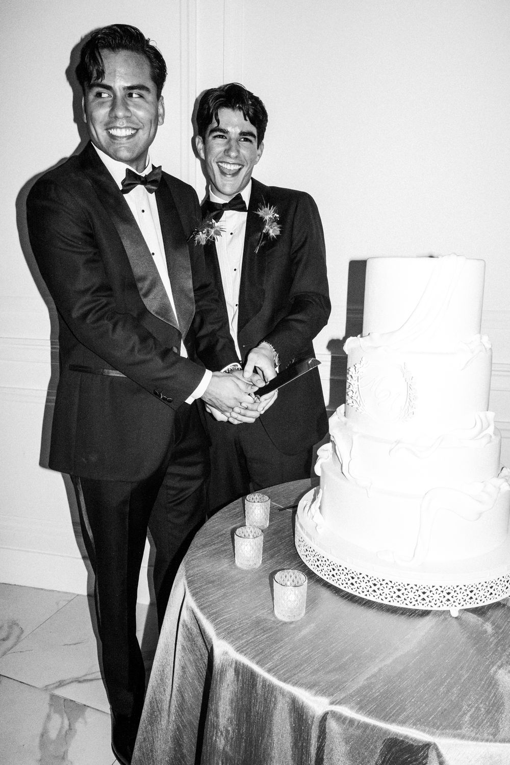 Two people in tuxedos cut an elegant white wedding cake together at a formal celebration.