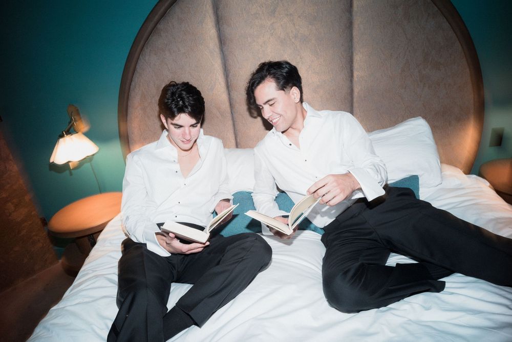 Two young men in white shirts and black trousers sit on a bed reading books in a stylish teal bedroom.
