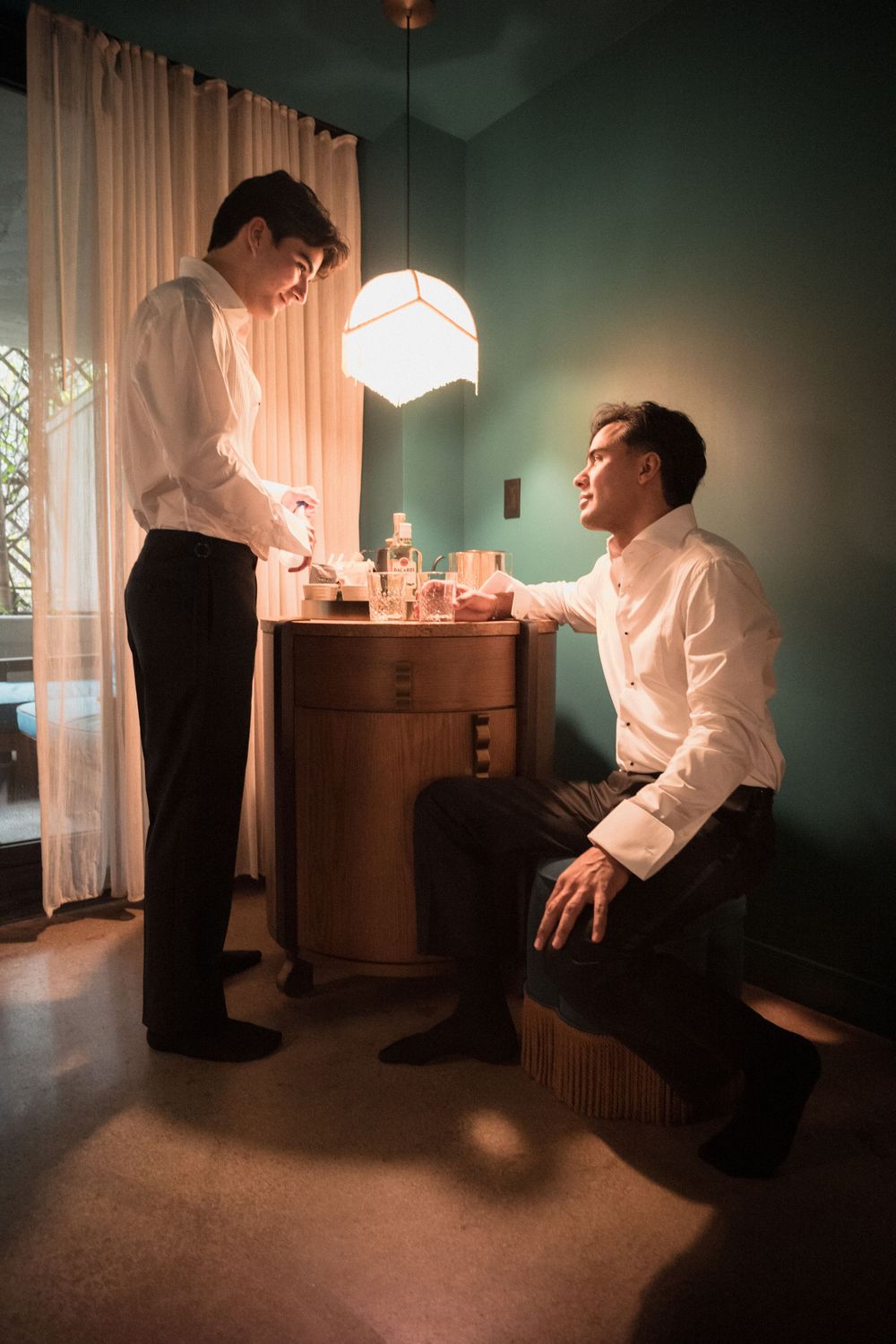 Two men in white dress shirts prepare together in a teal-walled dressing room with warm pendant lighting.