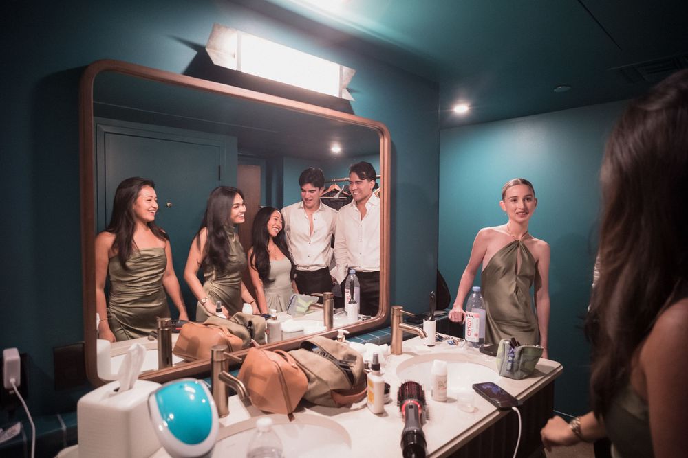 Group of friends laughing and socializing in a stylish teal dressing room with vanity mirror and makeup.