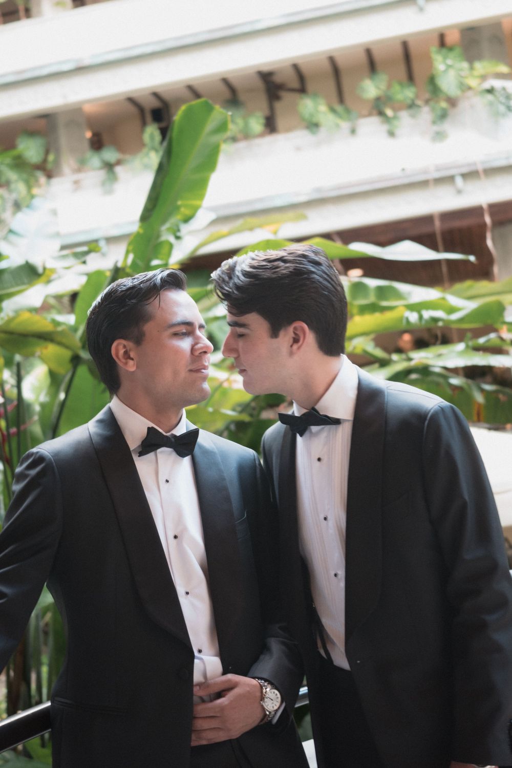 Two grooms in black tuxedos and bow ties share a romantic moment surrounded by tropical greenery.