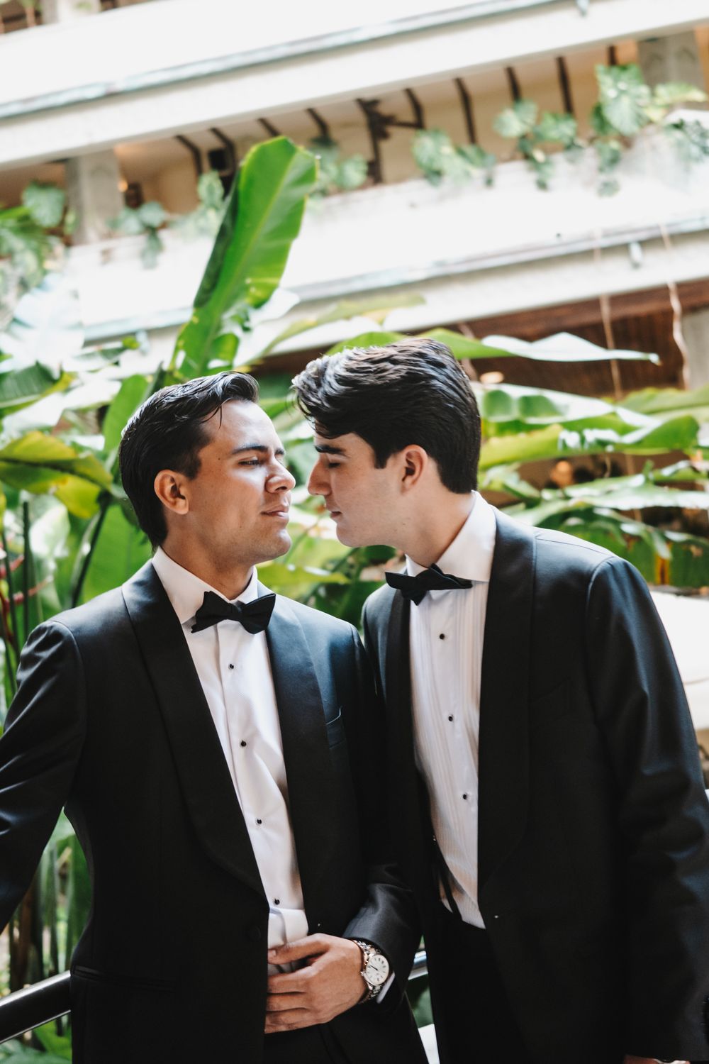 Two people in black tuxedos and bow ties having an intimate conversation in a garden setting.